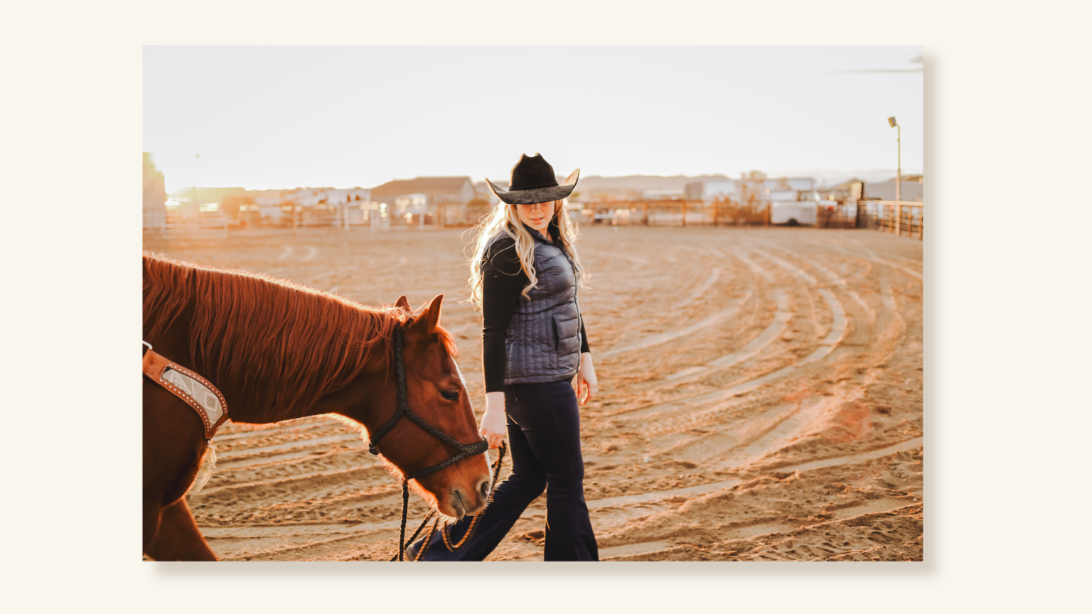 Woman in a cowboy hat leading a horse across a sandy arena at sunset, looking back toward the horse.